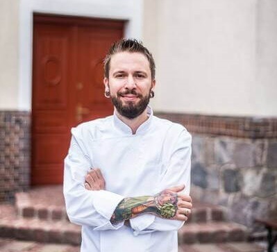 Szef kuchni w białym kitlu z tatuażami na przedramionach, ze skrzyżowanymi rękami, uśmiechnięty przy wejściu do restauracji, kamienne schody i drewniane drzwi.