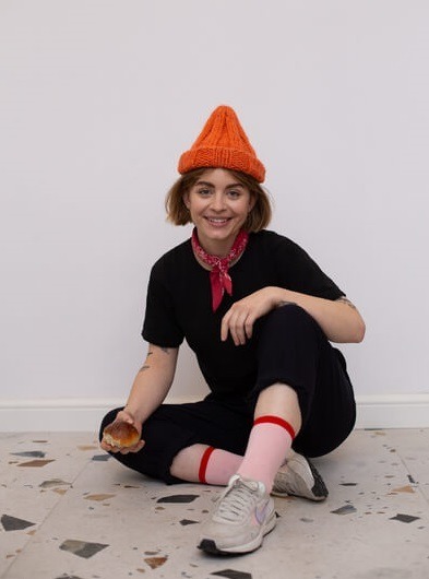 Uśmiechnięta młoda kobieta siedzi na posadzce z lastryko, w pomarańczowej czapce, czarnym ubraniu i różowych skarpetkach, trzyma bułkę.