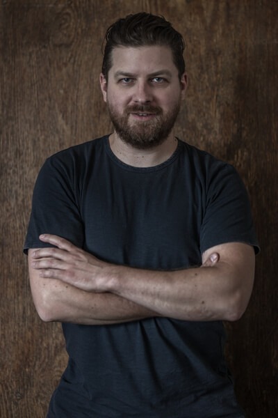 Brodaty mężczyzna w ciemnej koszulce, ramiona skrzyżowane na tle ciepłego drewnianego panelu, spokojny, pewny wyraz twarzy