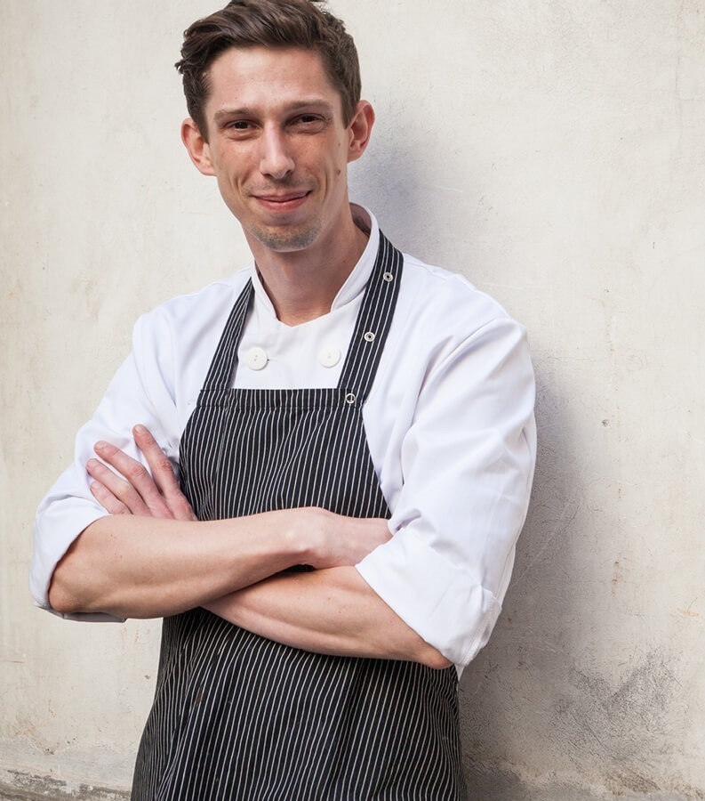 Uśmiechnięty szef kuchni hotelowej w białej kurtce i prążkowanym fartuchu, ręce skrzyżowane, oparty o fakturowaną ścianę