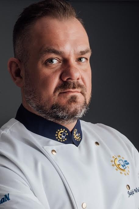 Szef kuchni w białym dwurzędowym mundurze z haftowanym herbem i granatowym kołnierzem, pewny wyraz twarzy, fine dining