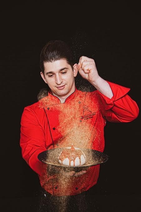 Szef w czerwonym kitlu sypiący cukrem pudrem na podany na talerzu deser — efektowna prezentacja deseru w restauracji