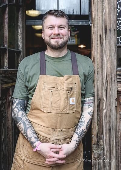 Uśmiechnięty kucharz w beżowym fartuchu z tatuażami stoi w rustykalnych drzwiach przytulnej restauracji