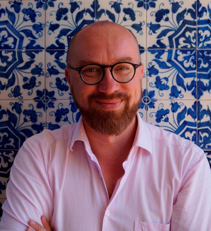 Uśmiechnięty mężczyzna z brodą i okrągłymi okularami w różowej koszuli na tle niebieskich, ręcznie malowanych płytek.