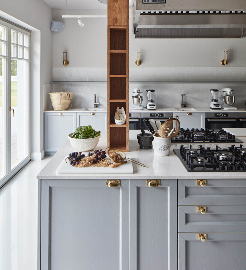 Jasna kuchnia z dużą szarą wyspą, płytą gazową, mosiężnymi uchwytami, drewnianą półką, robotami kuchennymi i misą z owocami i sałatą.