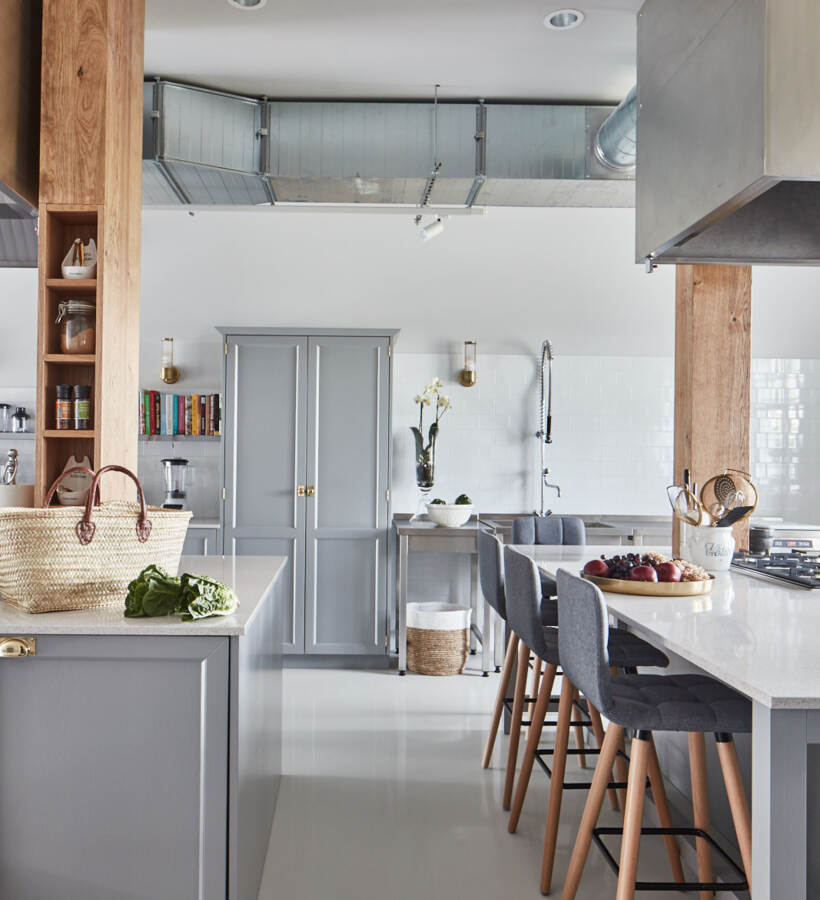 Jasna nowoczesna kuchnia z szarą wyspą i marmurowym blatem, czterema szarymi hokerami, odsłoniętym kanałem i drewnianymi belkami.