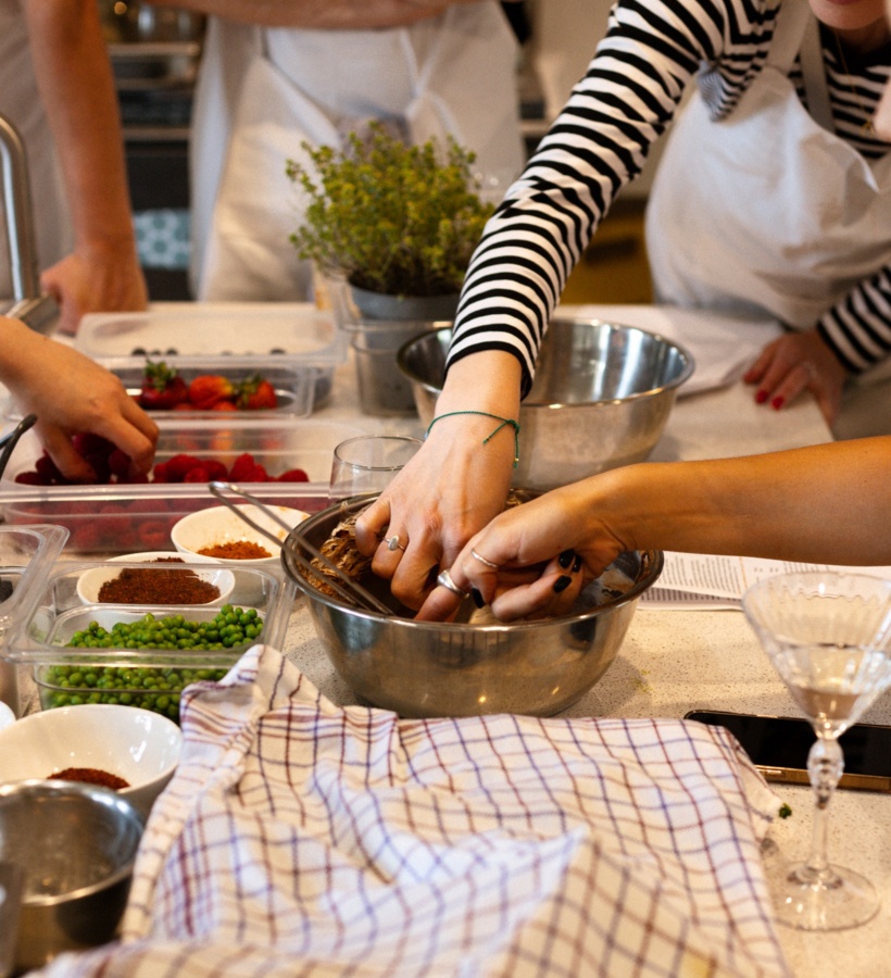 Goście w fartuchach na warsztatach kulinarnych mieszają ciasto w misce, obok pojemniki z groszkiem, truskawkami i ziołami.