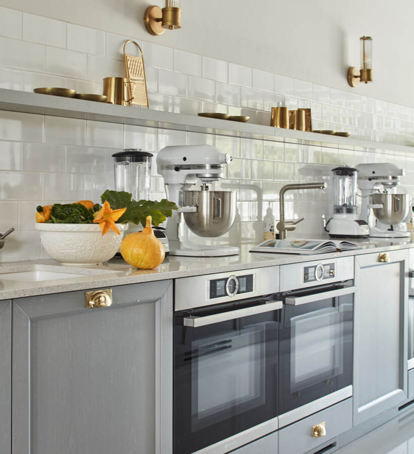 Jasna kuchnia hotelowa z szarymi szafkami, podwójnymi piekarnikami, białym blatem, robotami i złotymi kinkietami oraz miską świeżych warzyw
