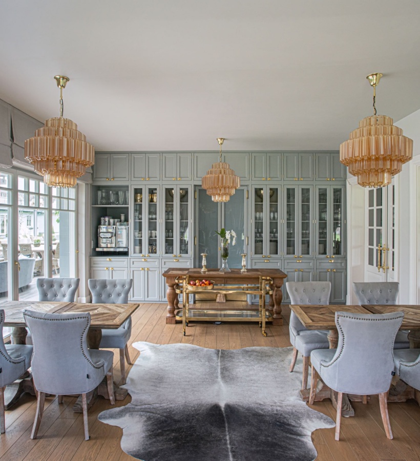 Elegancka jadalnia z pikowanymi szarymi krzesłami, drewnianymi stołami, przeszklonymi szafkami, mosiężnymi żyrandolami i dywanem ze skóry