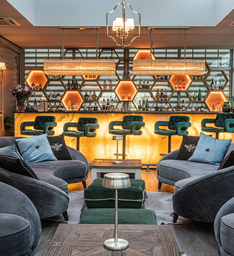 Hotelowy lounge z podświetlanymi sześciokątnymi półkami, barem z onyksu, zielonymi hokerami i szarymi sofami przy dużych oknach