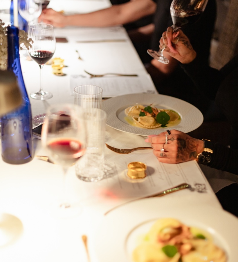 Długi stół z białym obrusem, ravioli na talerzach, goście trzymają kieliszki czerwonego wina, ciepła nastrojowa atmosfera