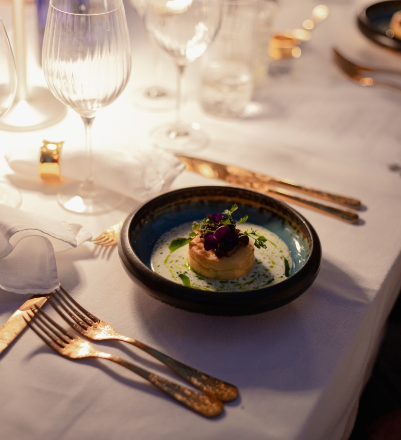 Elegancki stół restauracyjny przy świecach, złote sztućce, kieliszki i podana przekąska w ceramicznej miseczce, ozdobiona jadalnymi kwiatami.