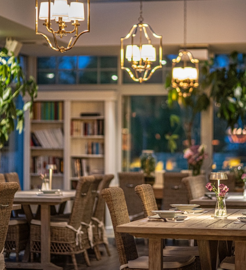 Przytulna restauracja hotelowa z drewnianymi stołami nakrytymi do kolacji, rattanowymi krzesłami, ciepłym oświetleniem i wazonikami przy oknach.