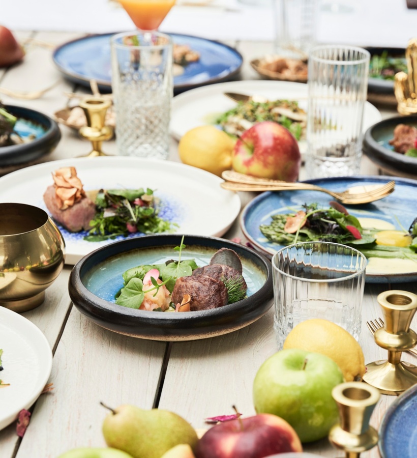Wspólne degustacyjne dania na niebieskiej ceramice, świeże owoce, kryształowe szklanki i mosiężne świeczniki na stole