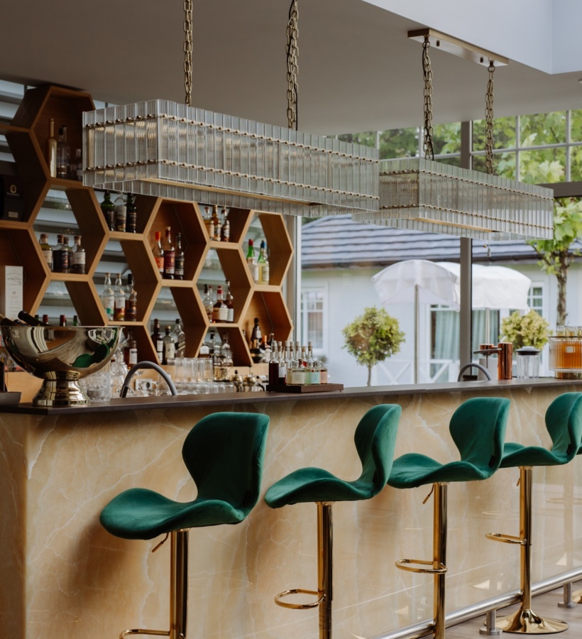 Bar hotelowy z zielonymi aksamitnymi hokerami, marmurowym blatem, sześciokątnymi półkami na alkohole i szklanymi lampami, widok na ogród.