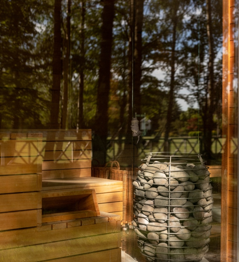 Drewniana sauna z przeszkloną ścianą, ławkami i metalowym piecem wypełnionym kamieniami oraz widokiem na las.