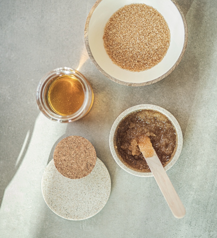 Miska z grubym brązowym cukrem, słoik złotego oleju i miseczka z peelingiem oraz drewniana szpatułka — naturalny zestaw spa.