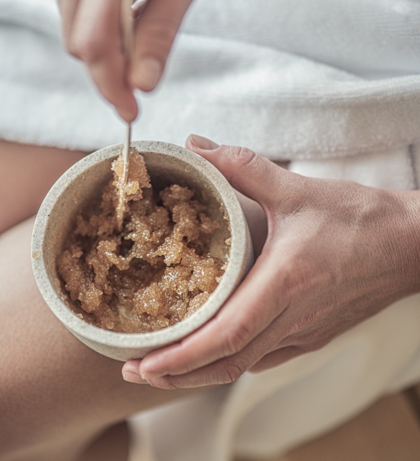 Ręce nabierające brązowego peelingu cukrowego z kamiennej miseczki, gość w białym szlafroku przed zabiegiem spa