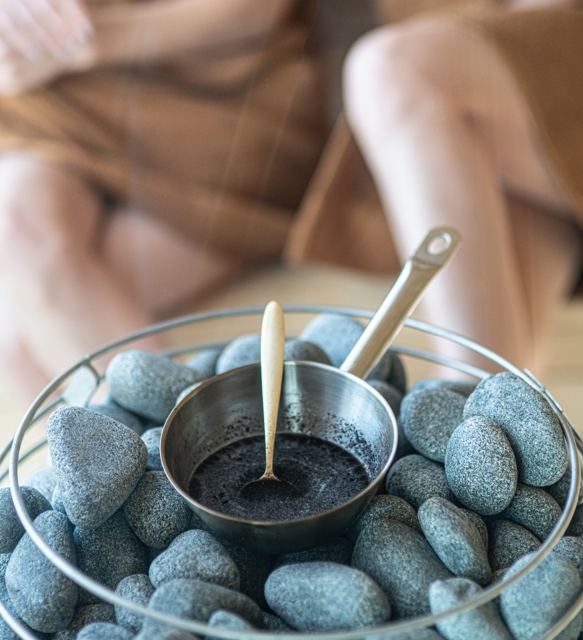 Metalowa miseczka z ciemnym peelingiem i łyżką na gładkich kamieniach sauny, w tle rozmyte osoby w szlafrokach