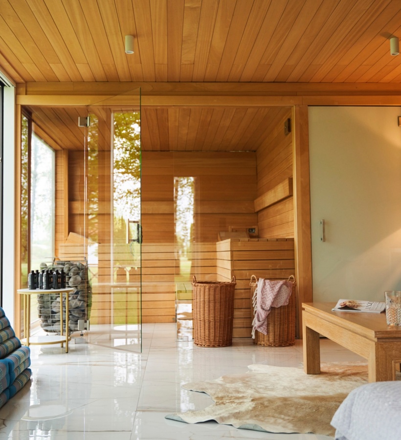 Sauna z drewnianym wnętrzem i przeszklonymi drzwiami obok przytulnego salonu, widok na las, wiklinowe kosze i herbata.