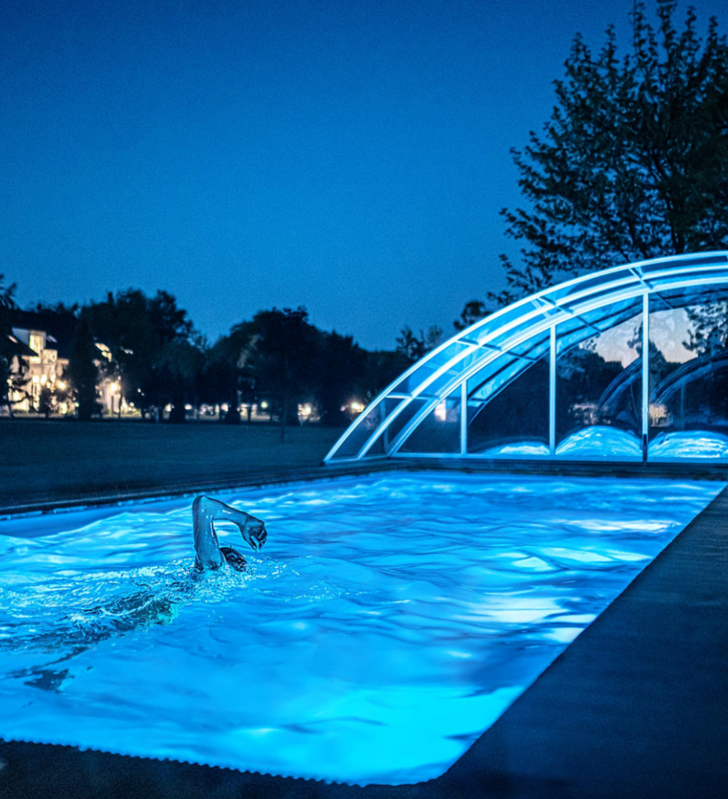Nocny basen na zewnątrz z niebieskim oświetleniem, gość płynie kraulem przy zakrzywionej, przesuwnej szklanej kopule, leżak i oświetlony hotel w tle.