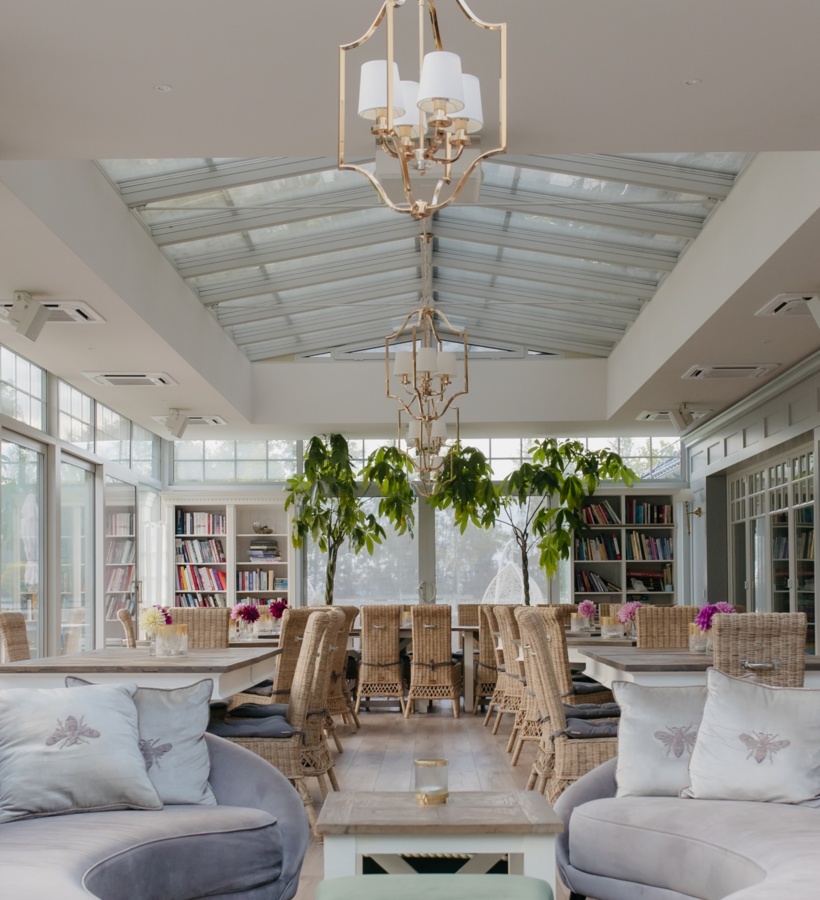Jasna oranżeria z przeszklonym dachem, złotymi żyrandolami, wiklinowymi krzesłami, zielonymi drzewkami, miękkimi sofami i regałami.