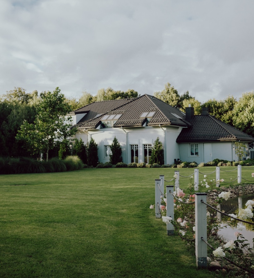 Willa wiejska z dachówką i świetlikami, zadbany trawnik, staw obsadzony kwiatami i spacerowa ścieżka z drewnianymi słupkami i liną