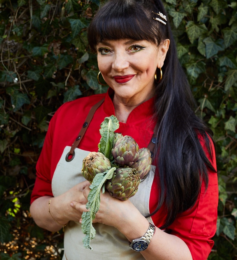 Uśmiechnięta szefowa kuchni w czerwonej marynarce i fartuchu trzyma świeże karczochy w ogrodzie, długie ciemne włosy i złote kolczyki.