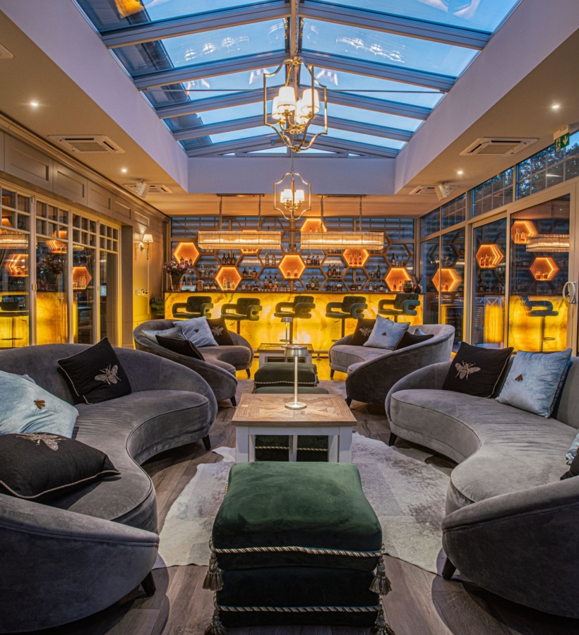 Luksusowy salon hotelowy z zaokrąglonymi aksamitnymi sofami, przeszklonym świetlikiem i podświetlanym sześciokątnym barem.