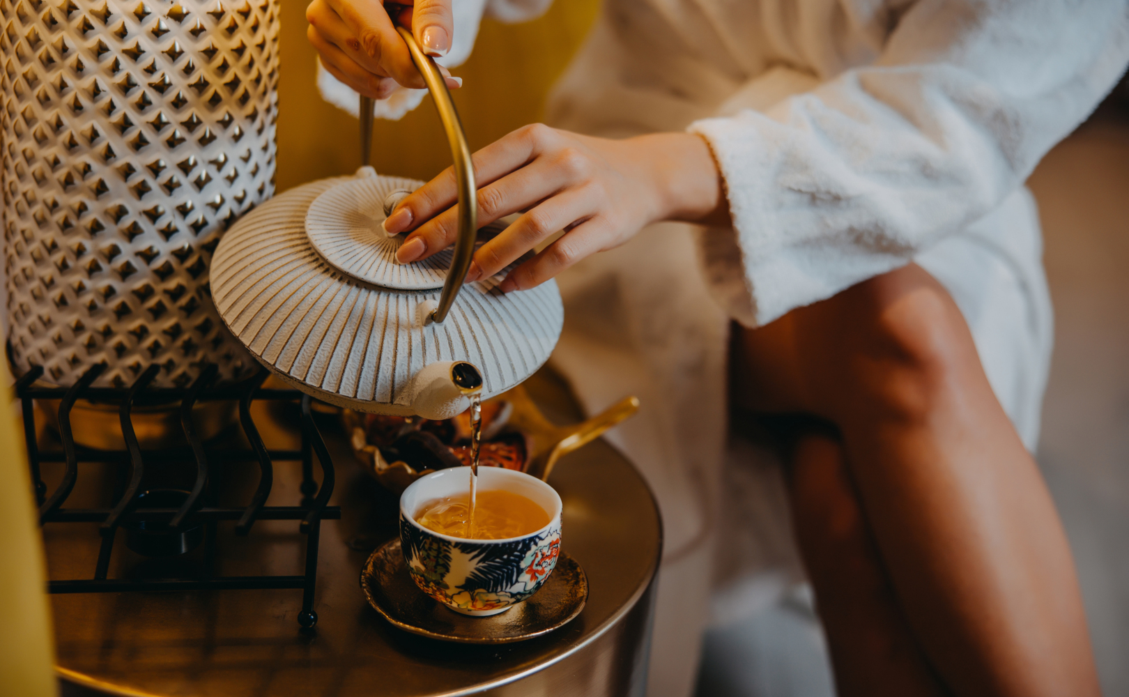 Host v bílém županu nalévá čaj z dekorativní konvičky do vzorovaného hrnku na kovovém stolku