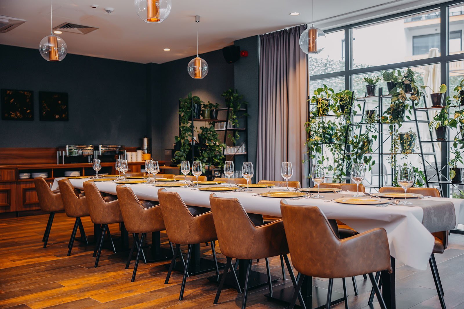 Nowoczesna jadalnia z długim stołem, skórzanymi krzesłami i szklankami, otoczona roślinami i dużymi oknami