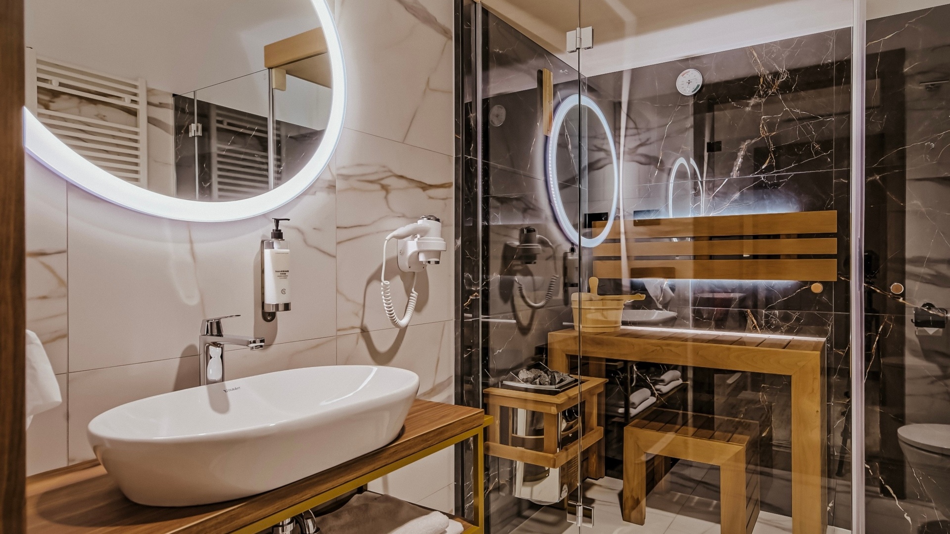 Łazienka hotelowa z nowoczesną umywalką, podświetlanym okrągłym lustrem oraz przeszkloną sauną z drewnianą ławką i akcesoriami.