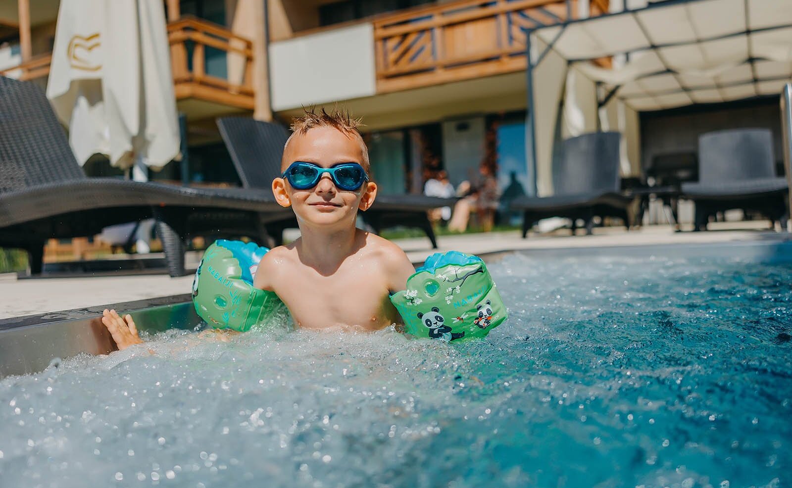 Mały chłopiec w okularach pływackich i zielonych rękawkach bawi się w basenie przy hotelu z leżakami i parasolem.