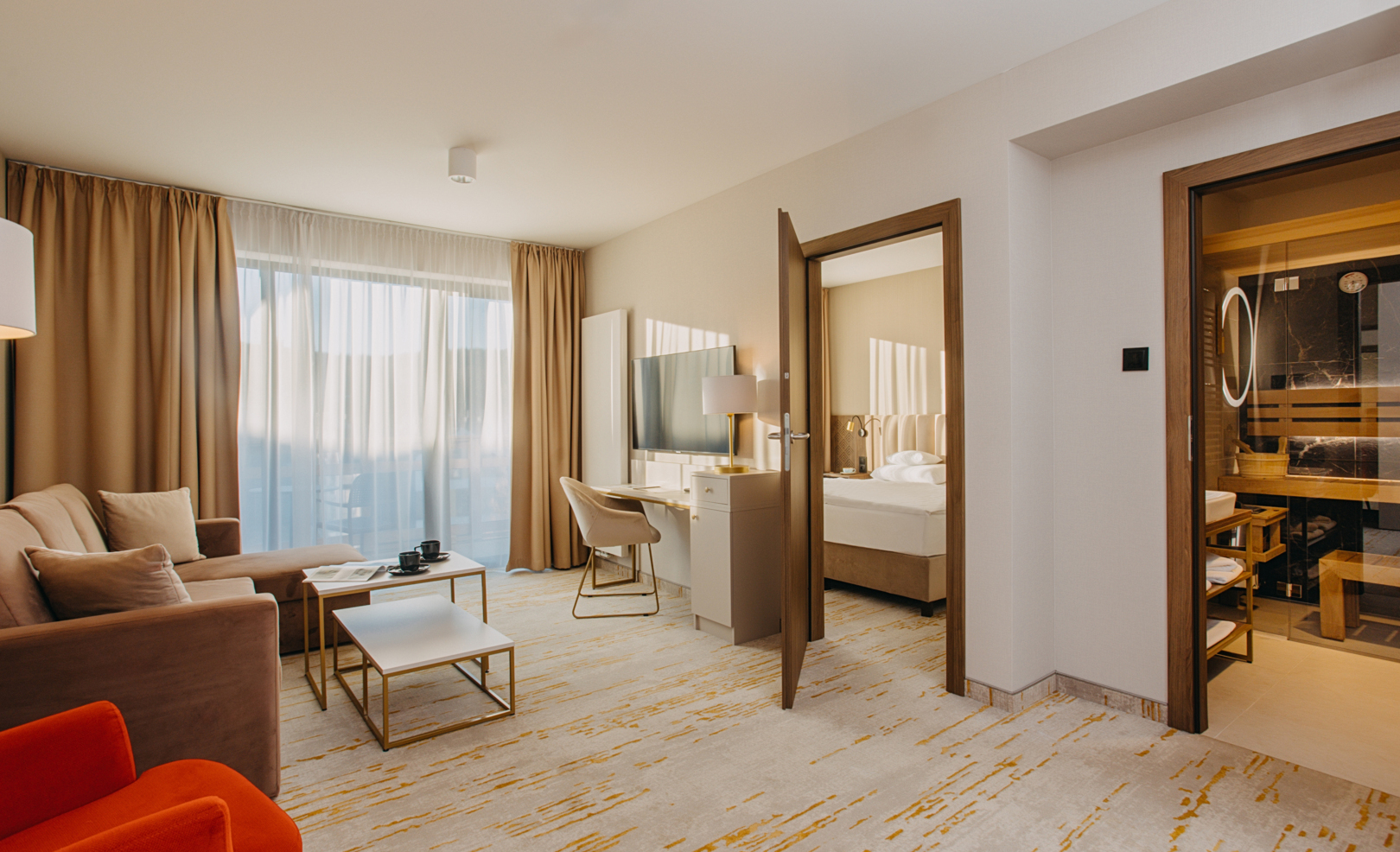 Großzügige Suite mit Wohnzimmer, Sofa, orangem Sessel, Schreibtisch, Schlafzimmer und Sauna neben großem Fenster.