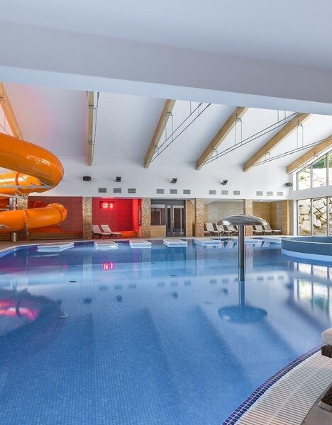 Moderní krytý hotelový bazén s oranžovou skluzavkou, lehátky a velkými skleněnými stěnami s výhledem na skalnatou zahradu