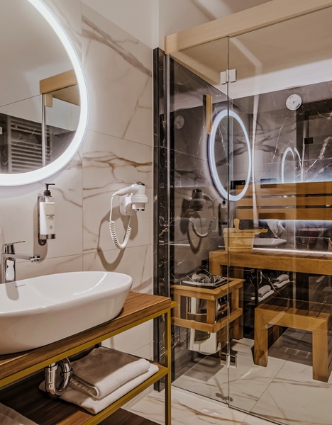Hotelová koupelna s moderním umyvadlem, osvětleným kulatým zrcadlem a prosklenou saunou s dřevěnou lavicí a doplňky.