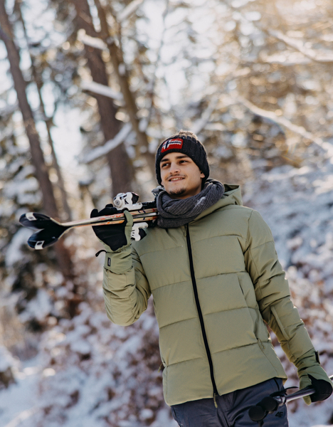 Mladý muž v zelené bundě a čepici nese lyže zimním zasněženým lesem.