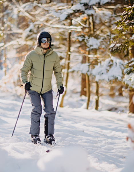 Osoba v zelené bundě a helmě běžkuje zasněženou lesní stezkou.
