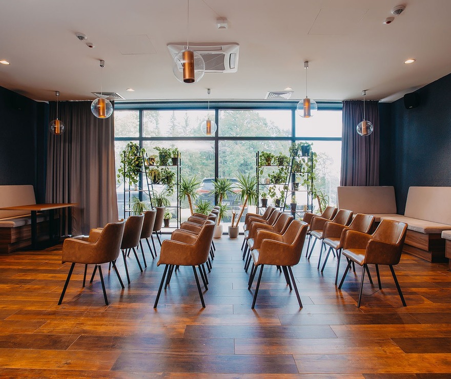 Nowoczesna sala konferencyjna z wygodnymi brązowymi krzesłami, dużymi oknami i zielonymi roślinami w dekoracyjnych stojakach.