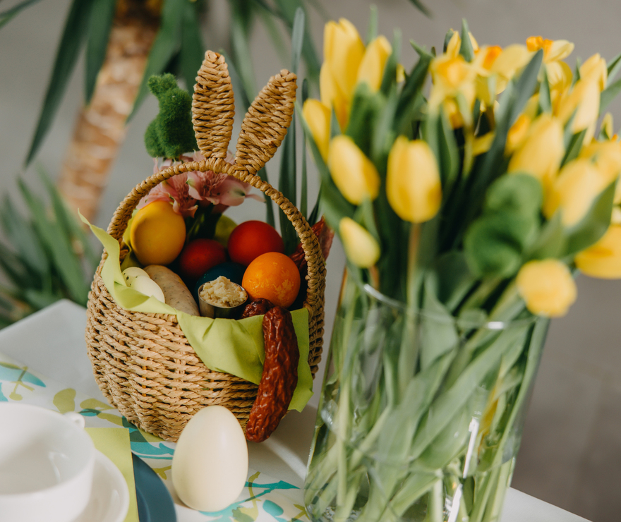 Wielkanocny koszyk ze święconką, kolorowe pisanki i wędliny na świątecznym stole obok żółtych tulipanów.