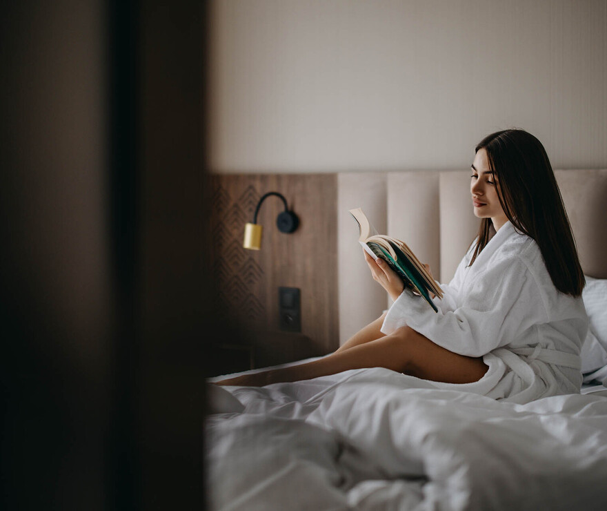Kobieta w białym szlafroku czyta książkę na wygodnym łóżku z miękkim zagłówkiem i jasną pościelą w nowoczesnym pokoju hotelowym.