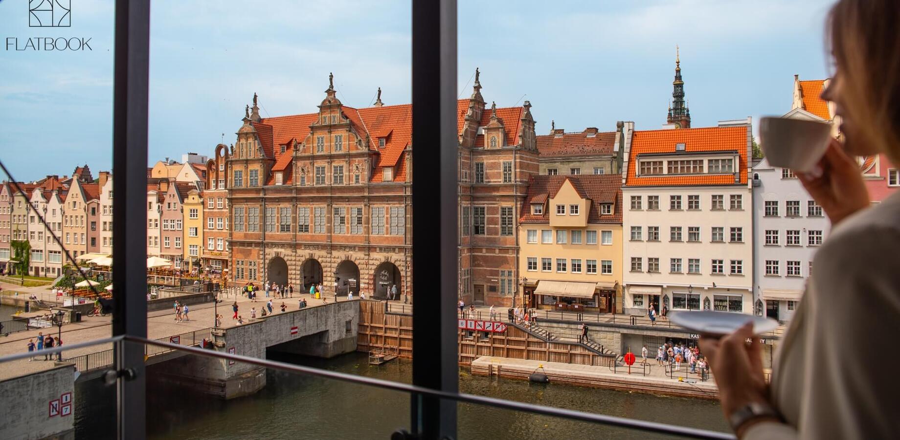 Gość z filiżanką na hotelowym balkonie, widok na zabytkową zabudowę z czerwonymi dachami nad kanałem i tętniący życiem deptak