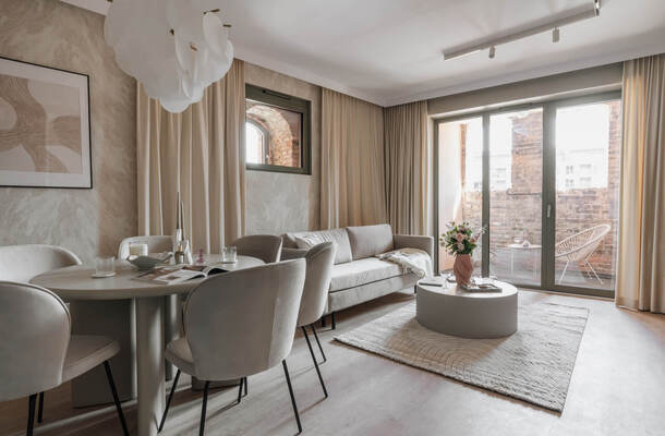 Przestronny, elegancki salon apartamentu z beżową sofą, jadalnią, dużymi przeszkleniami i prywatnym balkonem z ceglaną ścianą.
