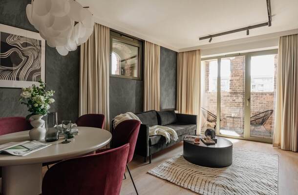 Salonik apartamentu z szarą sofą, okrągłym stołem i bordowymi krzesłami, balkonem z dwoma fotelami i widokiem na ceglany mur.