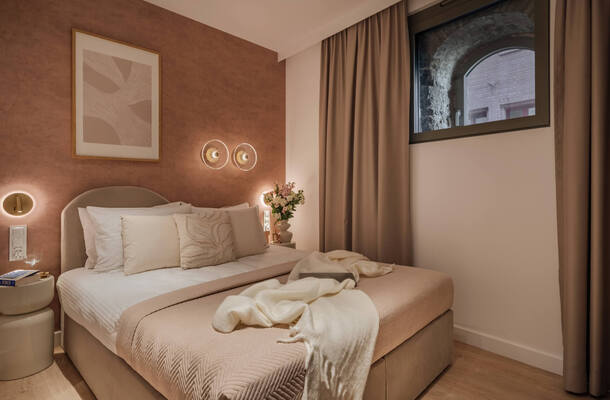 Przytulny pokój hotelowy w ciepłych beżowych tonach, łóżko z dekoracyjnymi poduszkami, nocne lampy, bukiet i łukowe okno.