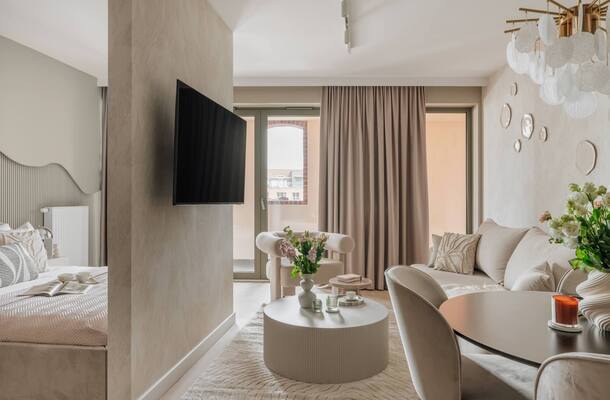 Przytulny apartament studio z wydzieloną sypialnią i salonem, telewizor na ściance, balkon, jasna sofa i kwiaty