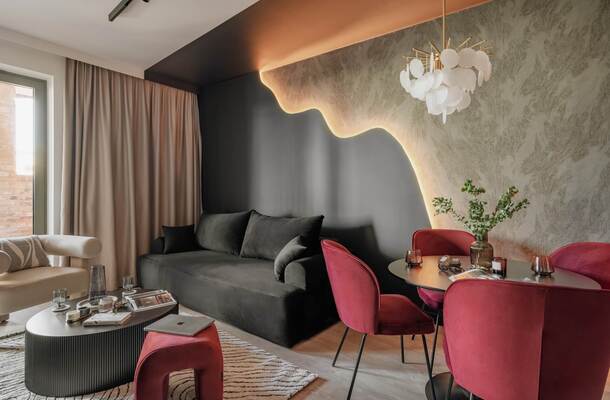 Stylowy apartament z ciemną sofą, kremowym fotelem, bordowymi krzesłami przy stoliku, podświetlanym dekoracyjnym panelem i eleganckim żyrandolem.