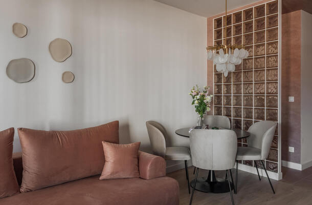 Kameralny lounge z terakotową sofą, okrągłym stolikiem i szarymi fotelami, szklaną ścianą dekoracyjną, ozdobną żyrandolową lampą i wazonem kwiatów.
