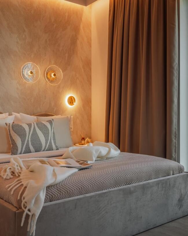 Przytulny pokój hotelowy z tapicerowanym łóżkiem, wzorzystą poduszką, ciepłym nastrojowym oświetleniem, zasłonami i miejskim widokiem przez okno.
