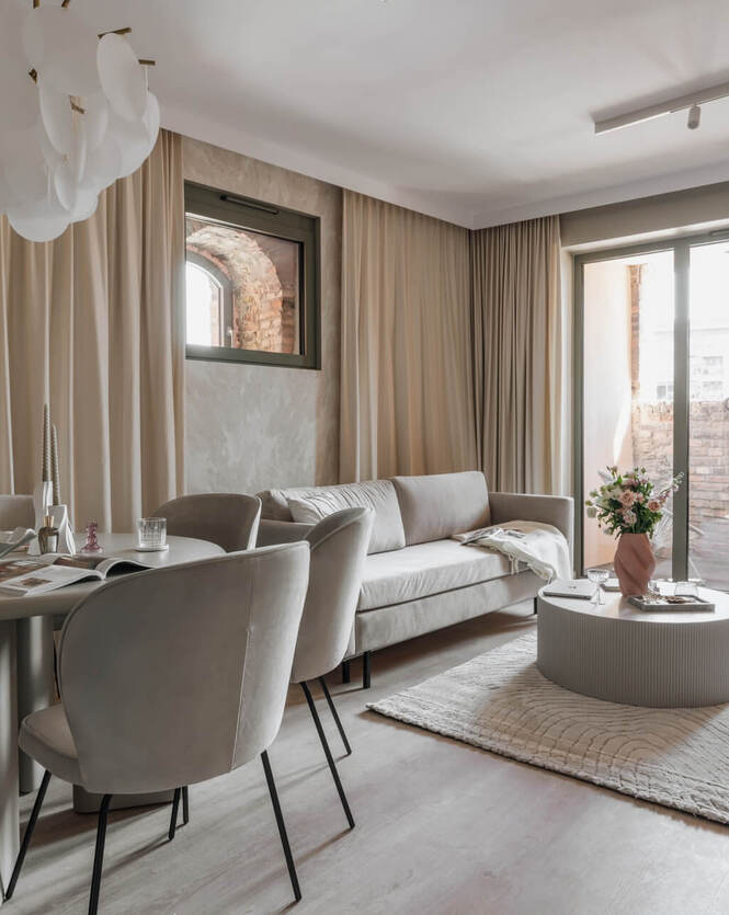 Przestronny, elegancki salon apartamentu z beżową sofą, jadalnią, dużymi przeszkleniami i prywatnym balkonem z ceglaną ścianą.
